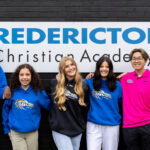 Grupo de estudantes internacionais sorrindo em frente à placa da Fredericton Christian Academy, escola particular cristã no Canadá que oferece programas de High School, Mini High School e Summer Camp com imersão acadêmica e cultural.