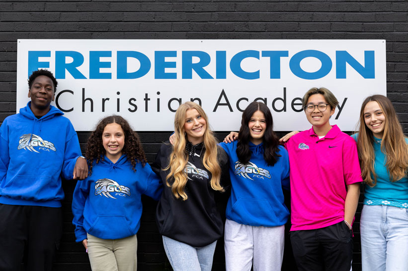 Grupo de estudantes internacionais sorrindo em frente à placa da Fredericton Christian Academy, escola particular cristã no Canadá que oferece programas de High School, Mini High School e Summer Camp com imersão acadêmica e cultural.