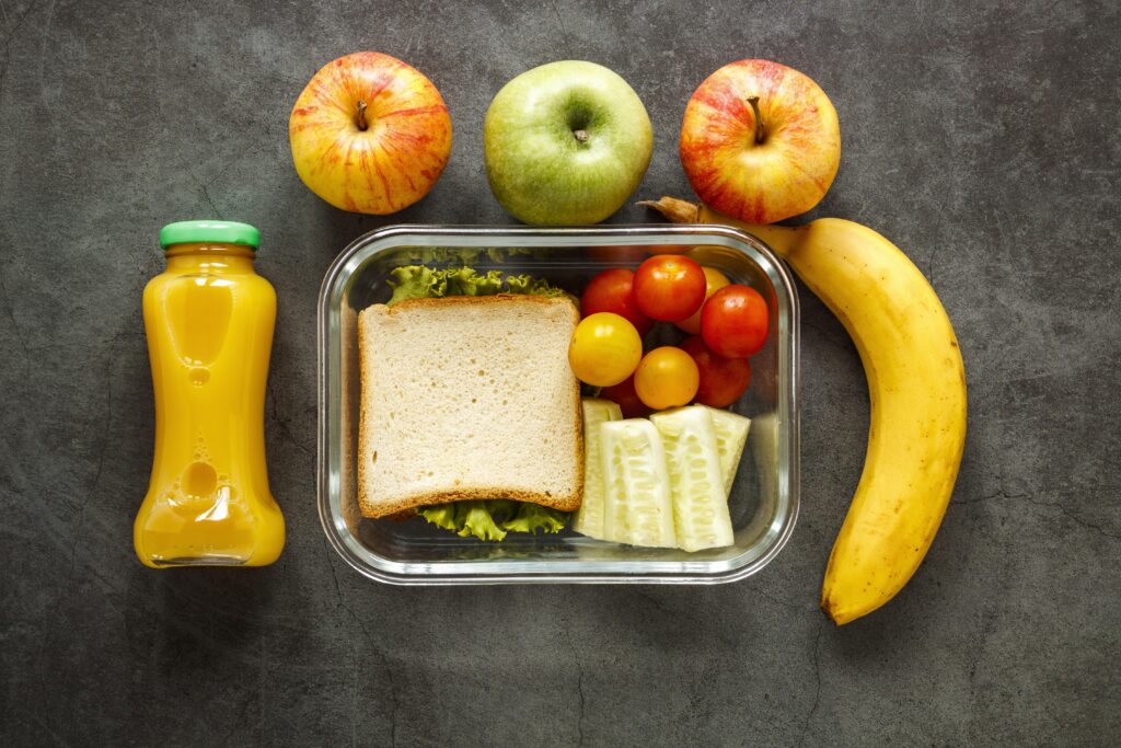 Marmita com sanduíche, frutas variadas e suco, representando uma das refeições diárias oferecida aos estudantes.