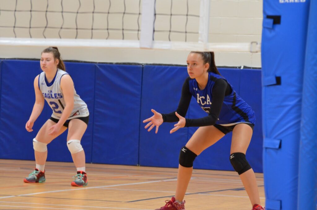 Estudantes da Fredericton Christian Academy em quadra de voleibol, prontas para receber o saque adversário.